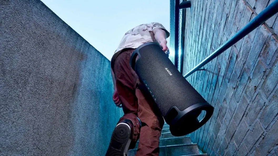 Person in Tarnshirt und brauner Hose trägt schwarzen Sony-Lautsprecher Treppe hinauf. Links dunkle Wand, rechts Ziegelwand mit Handlauf.