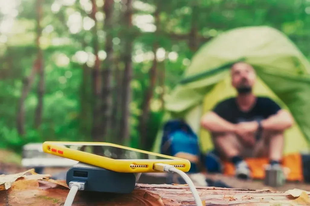 Gelbes Smartphone lädt im Campingzelt-Lager auf, Camper sitzt im Hintergrund.
