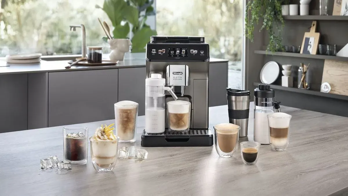 Grauer Holztisch mit Kaffeemaschine, verschiedenen Kaffeegläsern, Eiswürfeln, Milchschäumer. Im Hintergrund eine moderne Küche mit Schränken und Fenster.