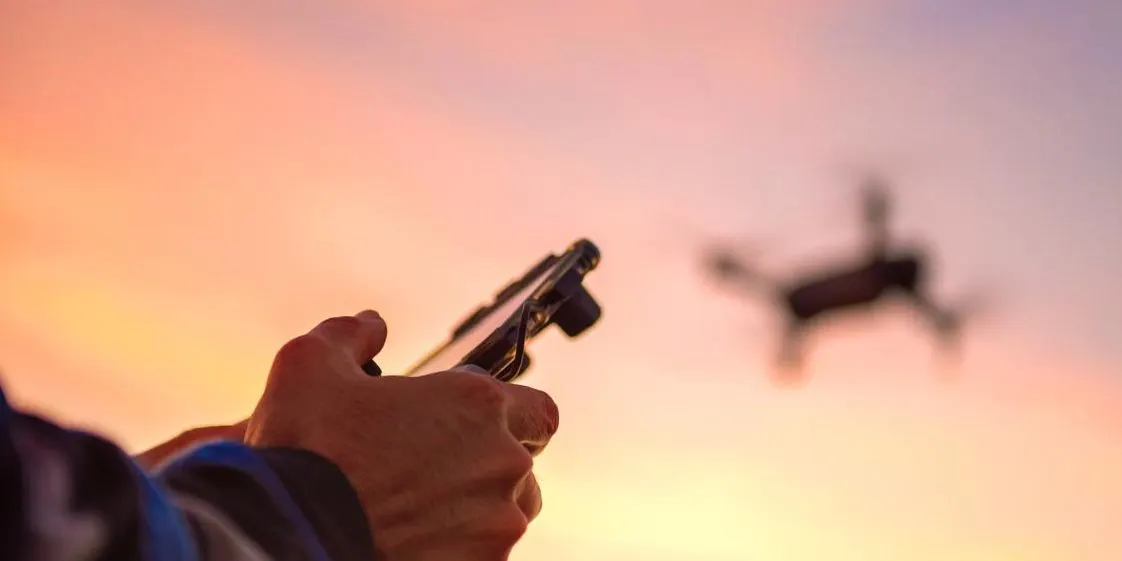 Person hält eine Fernbedienung in den Händen und steuert eine Drohne, die vor dem Abendhimmel fliegt.