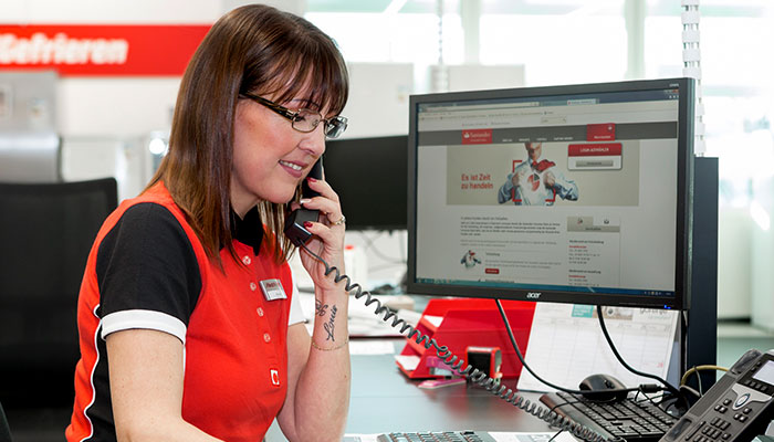 Eine Frau mit Brille telefoniert am Schreibtisch mit Computer und Telefon. Roter Schriftzug im Hintergrund.