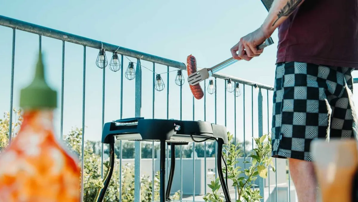 Eine Person grillt eine Wurst auf einem Balkon an einem Elektrogrill