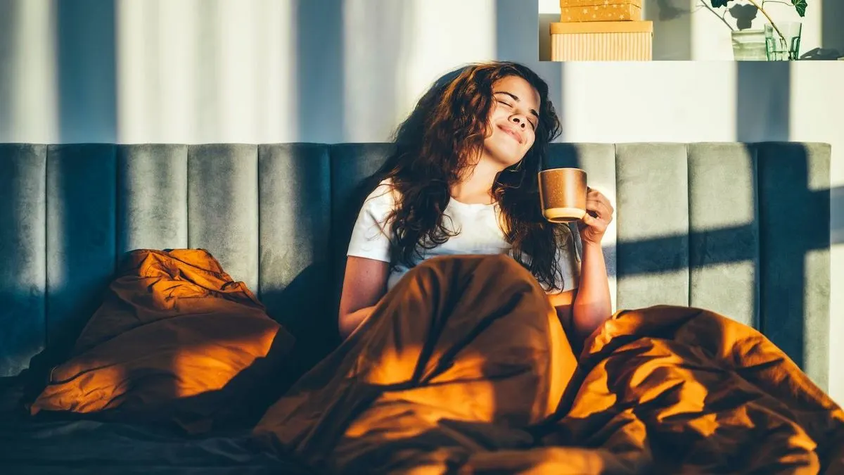 Person im Bett mit orangefarbener Decke, hält braune Tasse. Sonnenlicht fällt auf graues Kopfteil und Person