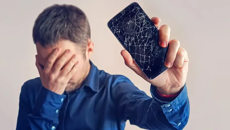 Person in blauem Hemd hält sich die Hand vors Gesicht und ein Smartphone mit stark gesprungenem Display hoch.