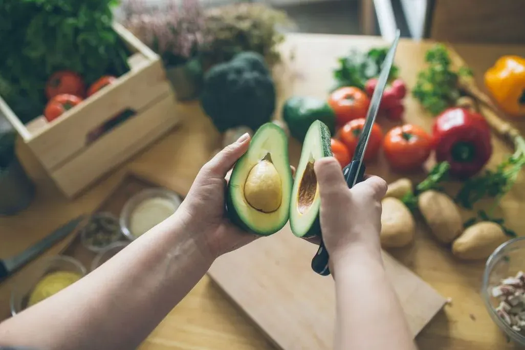 Hände halten eine aufgeschnittene Avocado mit Kern und ein Messer über einem Schneidebrett. Im Hintergrund sind Tomaten, Paprika und Brokkoli.