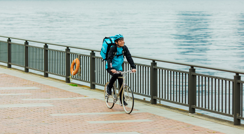 Ein Fahrradfahrer mit türkisfarbenem Rucksack fährt auf einem Weg neben einem See. Ein Rettungsring hängt an einem Geländer.