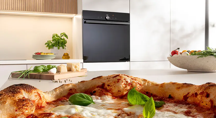 Eine Pizza mit Basilikum liegt auf einer Küchentheke vor einem schwarzen Ofen und Küchenutensilien.