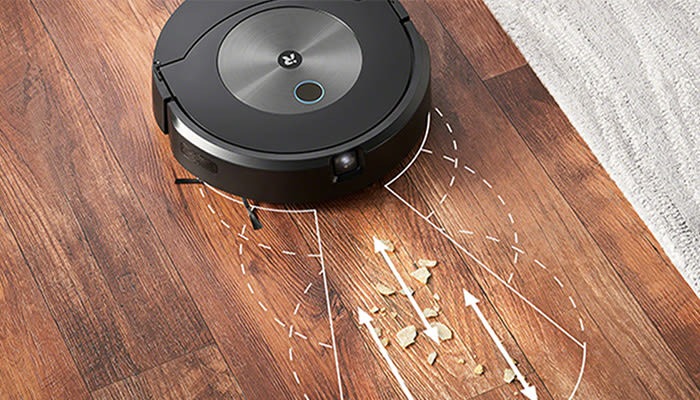 Ein schwarzer Saugroboter auf einem Holzboden mit Krümeln. Pfeile zeigen die Richtung der Reinigung an. Ein Teppich ist teilweise sichtbar.