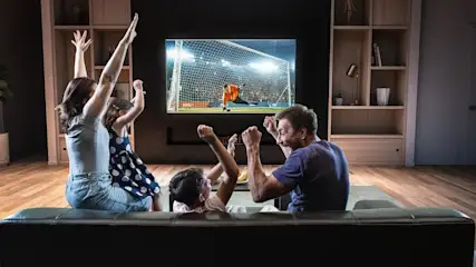 Familie jubelt auf schwarzem Sofa, schaut Fußballspiel mit Torwartparade auf großem TV. Im Hintergrund Holzregal und Holzboden.