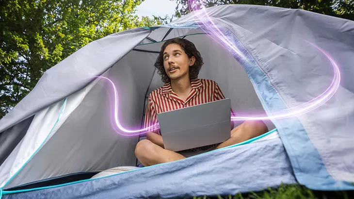 Mann sitzt mit einem Laptop in einem Zelt im Freien; leuchtende violette Linien symbolisieren digitale Vernetzung.