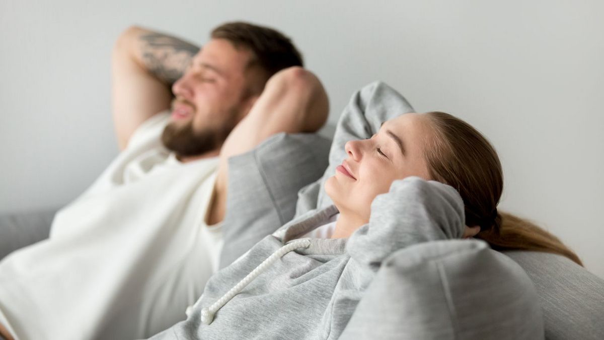 Zwei Personen sitzen entspannt nebeneinander auf einem Sofa und lehnen sich mit den Händen hinter dem Kopf zurück.