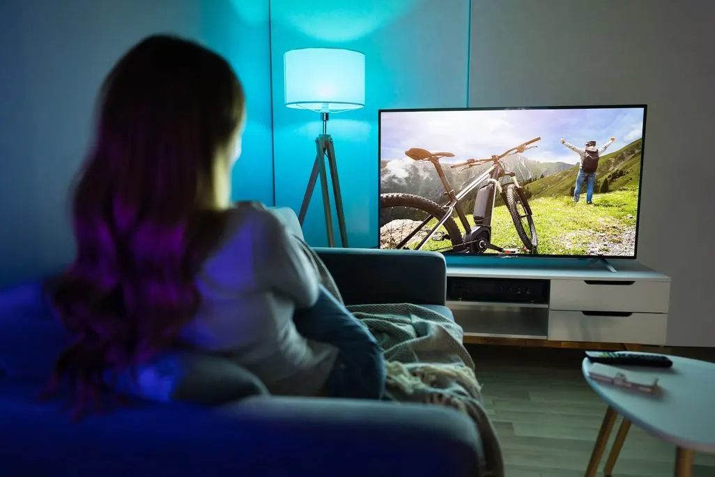Eine Frau sitzt auf einem Sofa und schaut auf einen Fernseher der eine Mountainbike-Szene zeigt.
