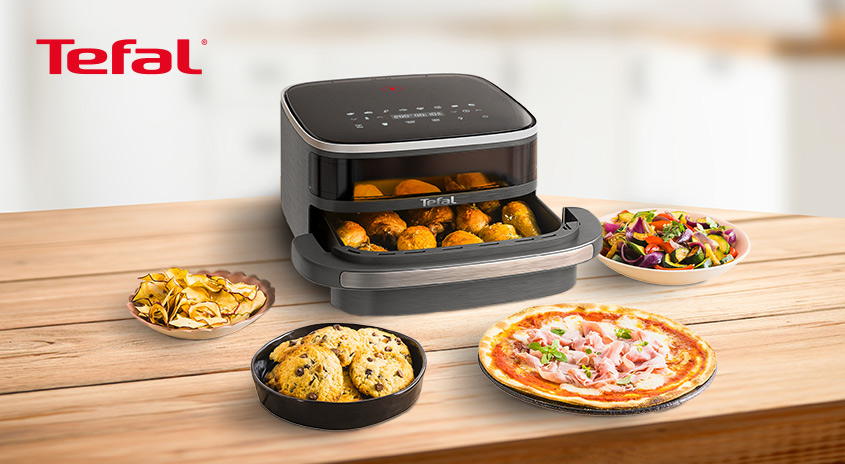 Auf einem Holztisch stehen eine Heißluftfritteuse, Pommes, Kekse, Pizza und Salat. Die Fritteuse ist mit Essen gefüllt.