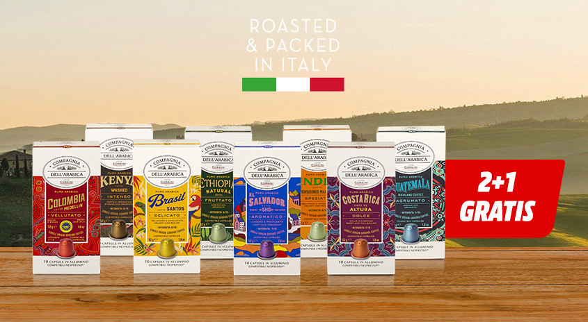 Kaffeekapseln in Kartons auf Holztisch vor Landschaft. Italienische Flagge und Text "2+1 GRATIS" im Hintergrund.