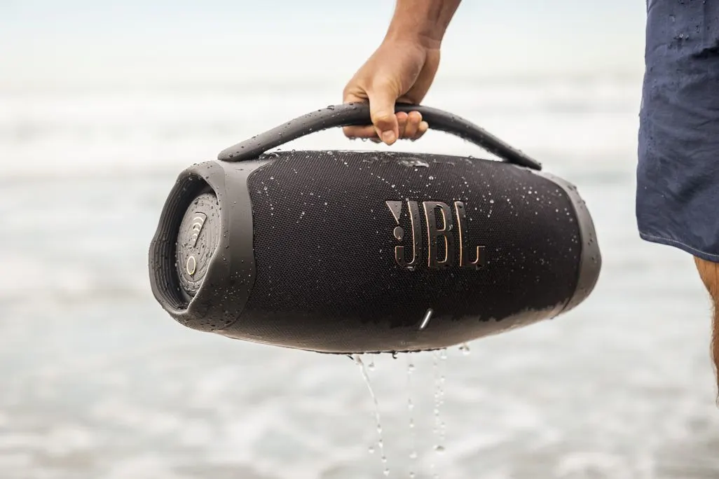 Eine Person hält eine nasse JBL Musikbox am Henkel am Strand.
