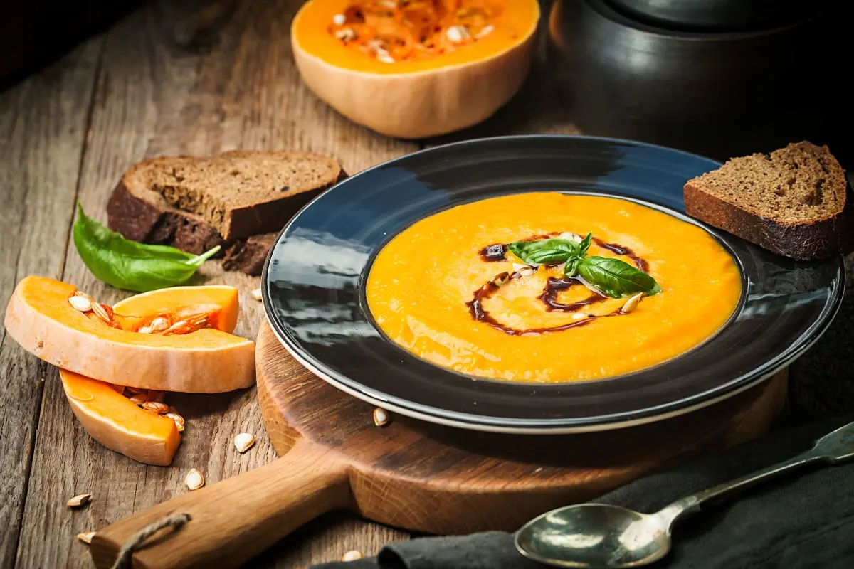 Kürbiscremsuppe in dunklem Teller, daneben liegt eine Scheibe Brot