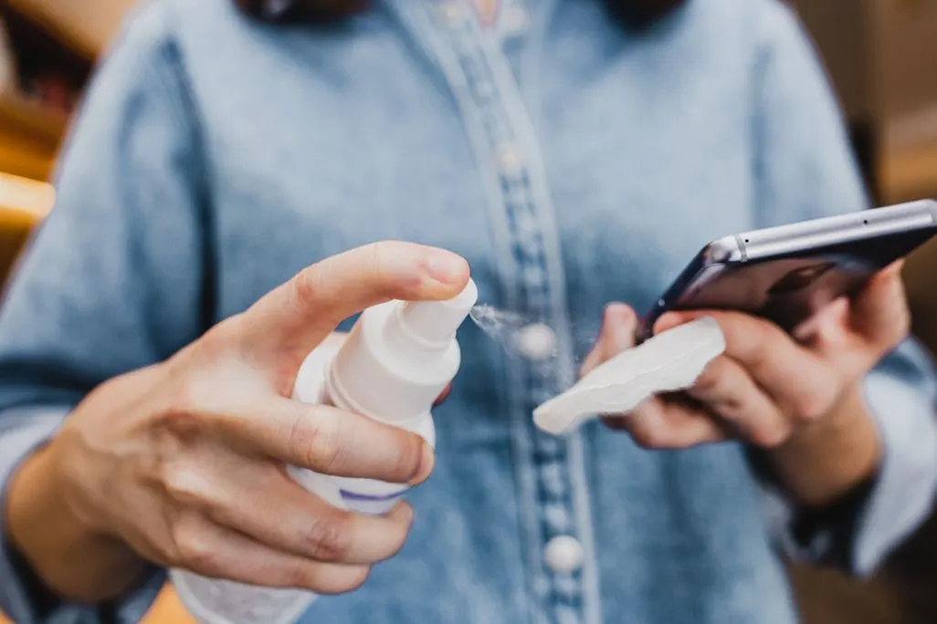 Person sprüht Reiniger auf ein Tuch und hält ein Smartphone