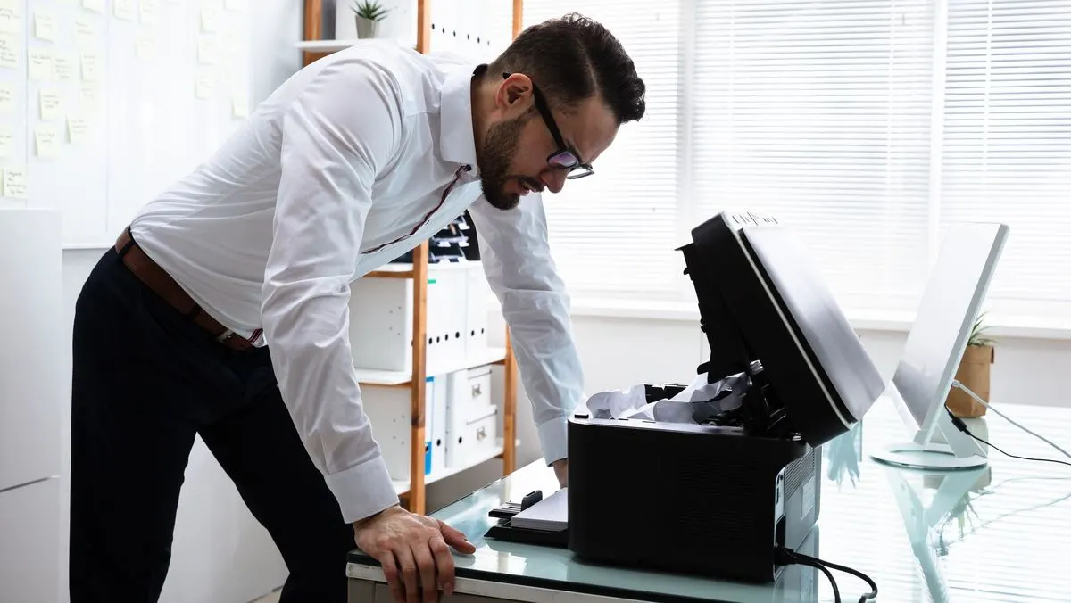 Ein Mann lehnt sich über einen Drucker im Buero und schaut in das Gerät, das mit zerknülltem Papier verstopft ist.