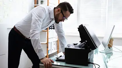 Ein Mann lehnt sich über einen Drucker im Buero und schaut in das Gerät, das mit zerknülltem Papier verstopft ist.