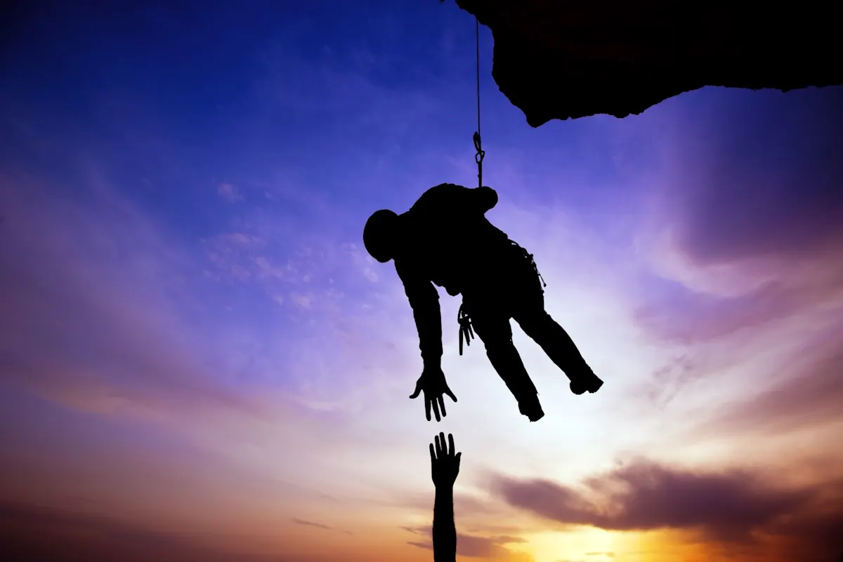 Die Silhouette eines Bergretters, der sich abseilt und versucht die hochgestreckte Hand eines Wanderers zu ergreifen.
