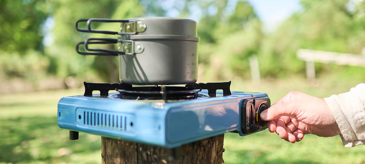 Auf einem Baumstumpf steht ein blauer Campingkocher mit einem Topf darauf. Eine Hand dreht den Knopf des Kochers.