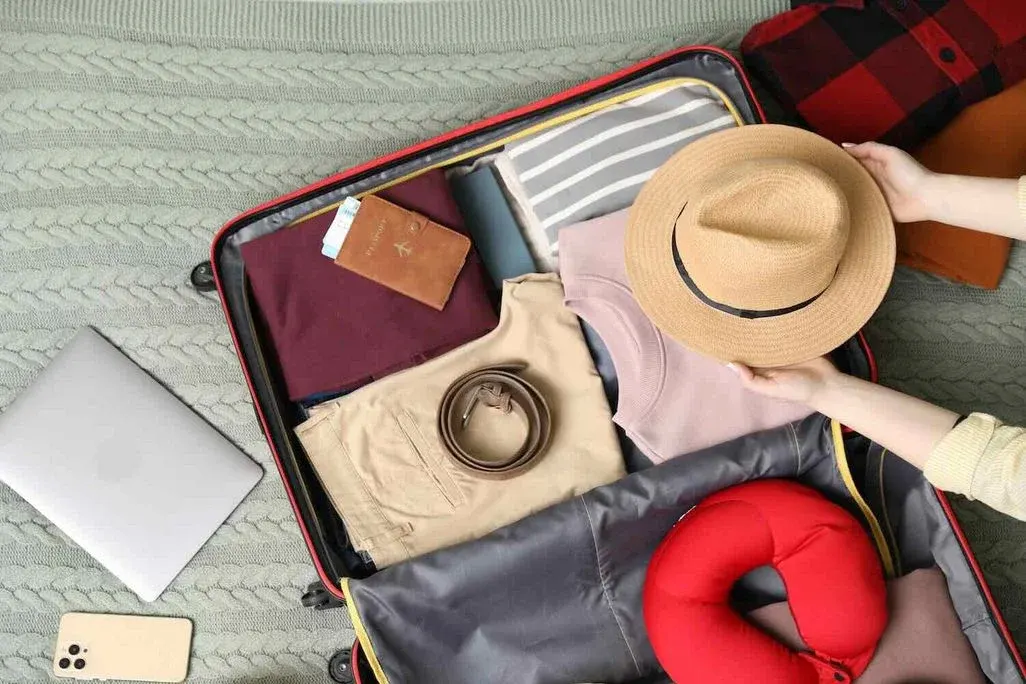 Hände halten einen Strohhut über einen fast fertig gepackten Koffer.