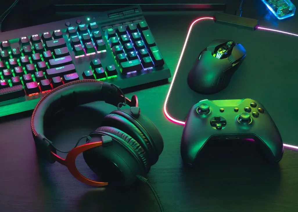 Schwarze Gaming-Tastatur mit lebendiger RGB-Beleuchtung, Headset mit orangefarbenen Akzenten, Maus auf leuchtendem Pad und Controller.