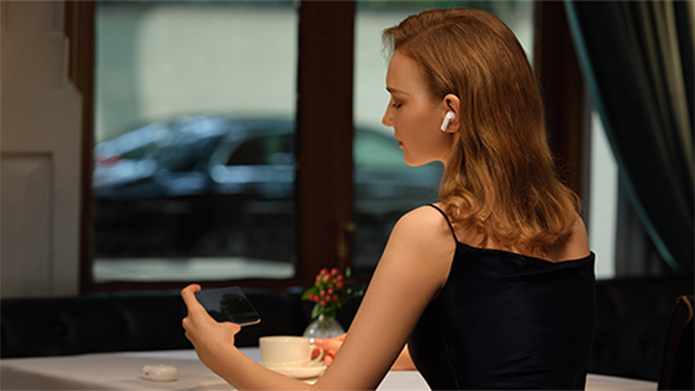 Eine Frau mit Kopfhörern hält ein schwarzes Smartphone an einem Tisch. Im Hintergrund ein Fenster und Vorhänge.