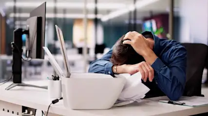 Frustrierter Mann sitzt am Schreibtisch, stützt den Kopf auf die Arme neben einem Drucker mit Papierstau.