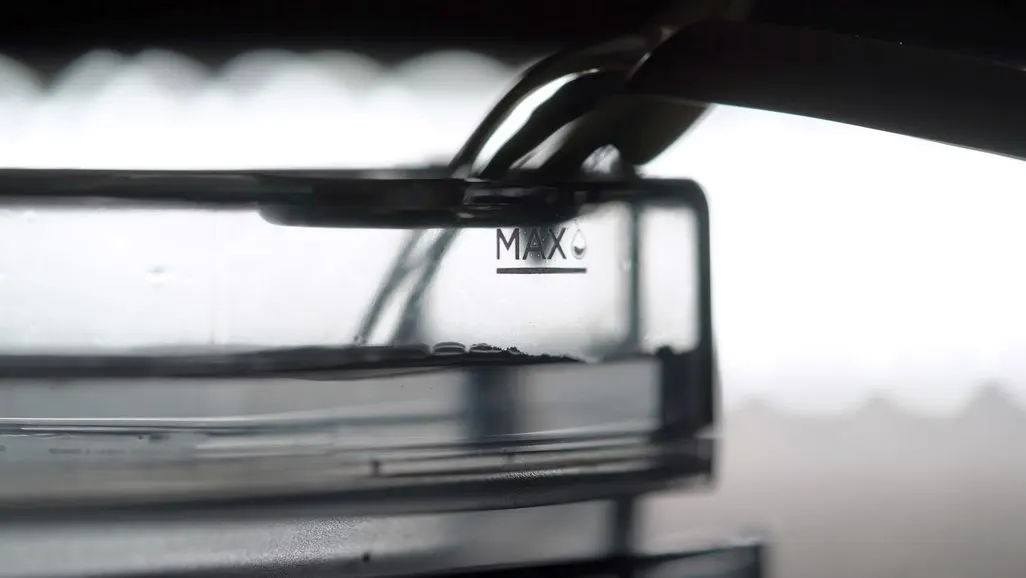 Wasser wird in einen transparenten Behälter gegossen und erreicht die "MAX"-Linie mit Tropfensymbol. Dunkler Ausguss sichtbar.