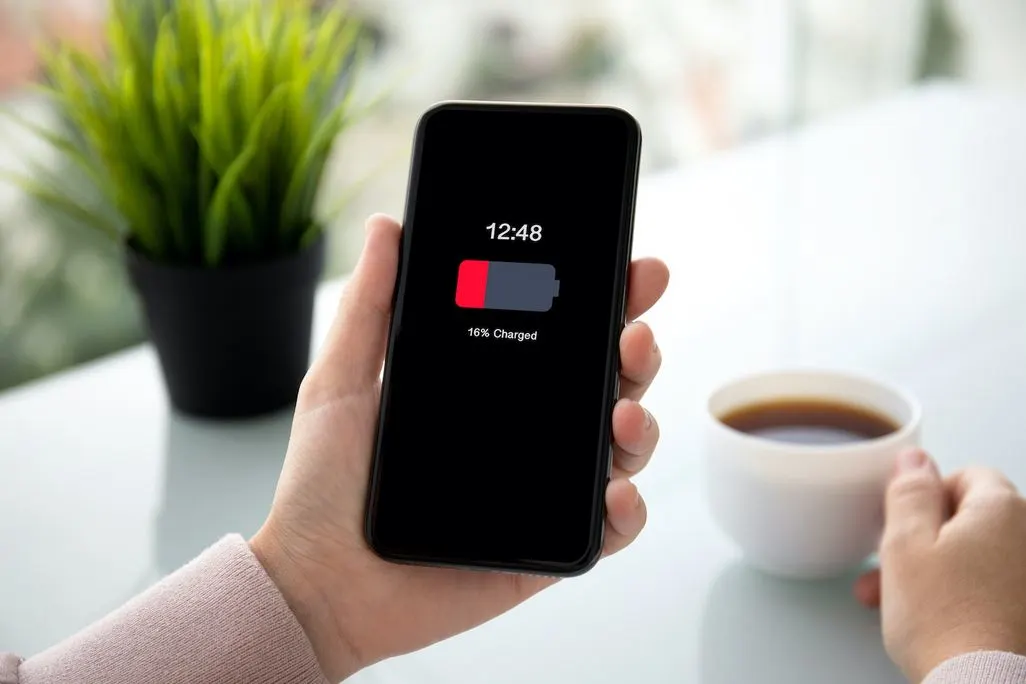 Hand hält ein Smartphone mit niedrigem Akkustand auf dem Bildschirm, im Hintergrund eine Tasse Kaffee.
