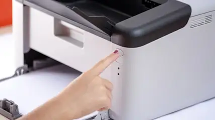 Eine Hand drückt den Einschaltknopf eines hellgrauen Druckers. Weißes Papier liegt im Fach, im Hintergrund ein orangefarbener Gegenstand.