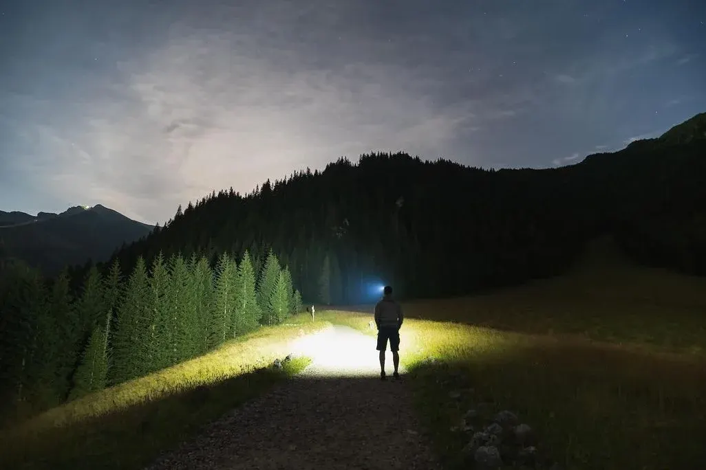 Nachtwanderer mit Stirnlampe auf Waldweg in Berglandschaft unter Sternenhimmel.