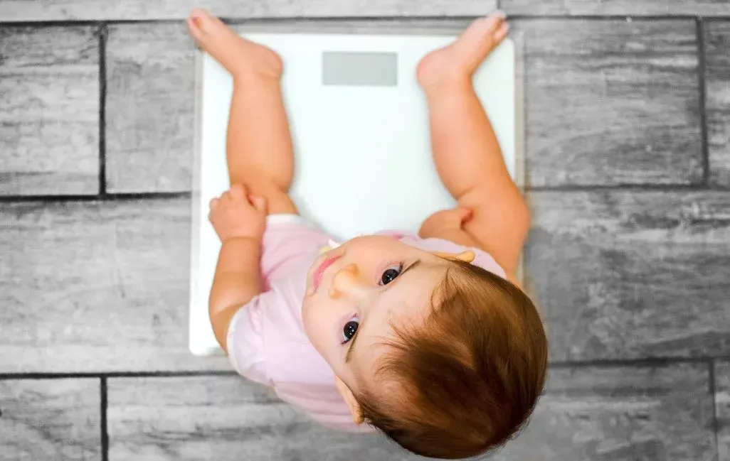 Baby in rosa Oberteil sitzt auf einer weißen Waage auf grauem Holzboden und blickt nach oben.