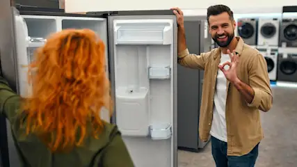 Ein Mann lächelt und zeigt ein OK-Zeichen neben einem geöffneten, leeren Kühlschrank. Eine Person blickt hinein. Im Hintergrund sind Waschmaschinen zu sehen.