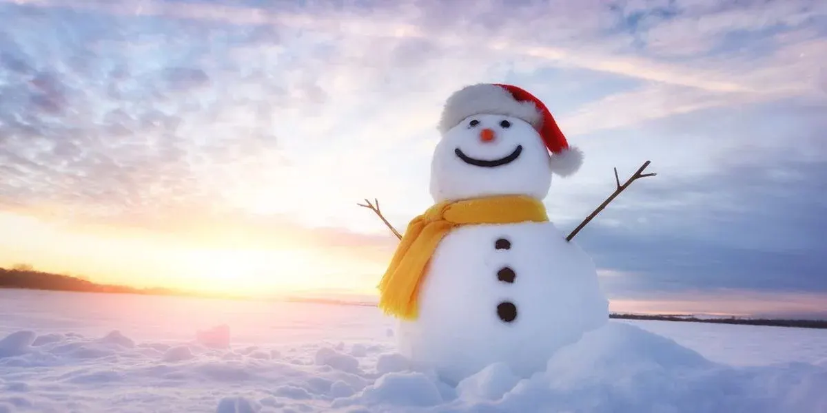 Schneemann mit lächelndem Gesicht, roter Mütze, gelbem Schal und Stockarmen auf Schnee. Orange-blauer Himmel bei Sonnenuntergang.