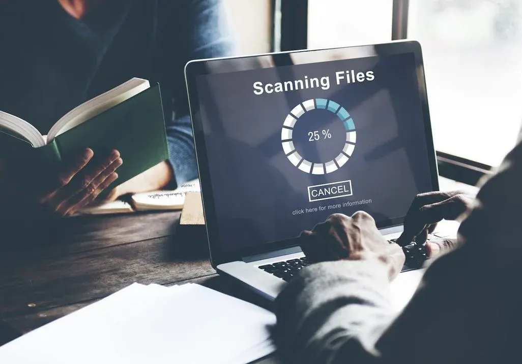 Person tippt auf Laptop; Bildschirm zeigt „Scanning Files“ mit Fortschrittskreis 25% und „Cancel“-Button, Bücher und Notizen im Hintergrund.