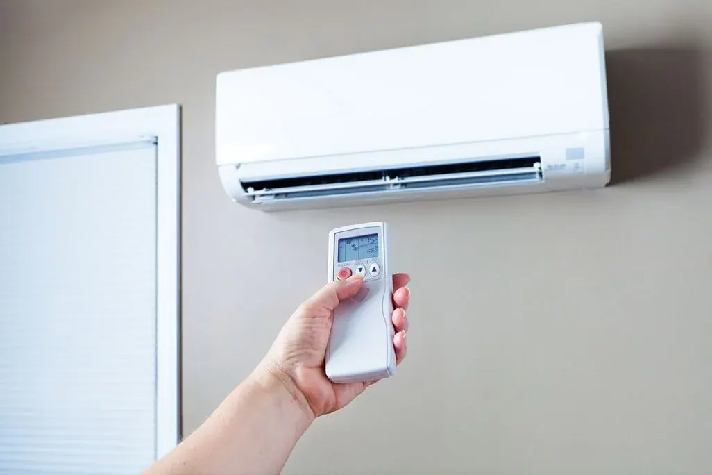 Hand richtet Fernbedienung auf eine weiße Klimaanlage an der Wand.