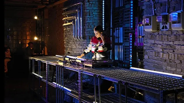 Eine Person in rot-weißem Oberteil steht an einem Tisch mit Apparaten vor einer Steinwand mit Rohren und blauen Lichtern.