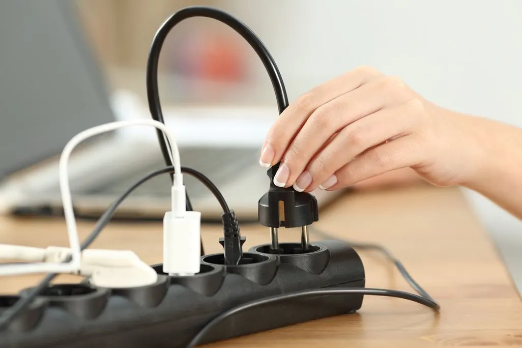 Hand steckt einen Stecker in Steckdosenleiste mit mehreren angeschlossenen Geräten.