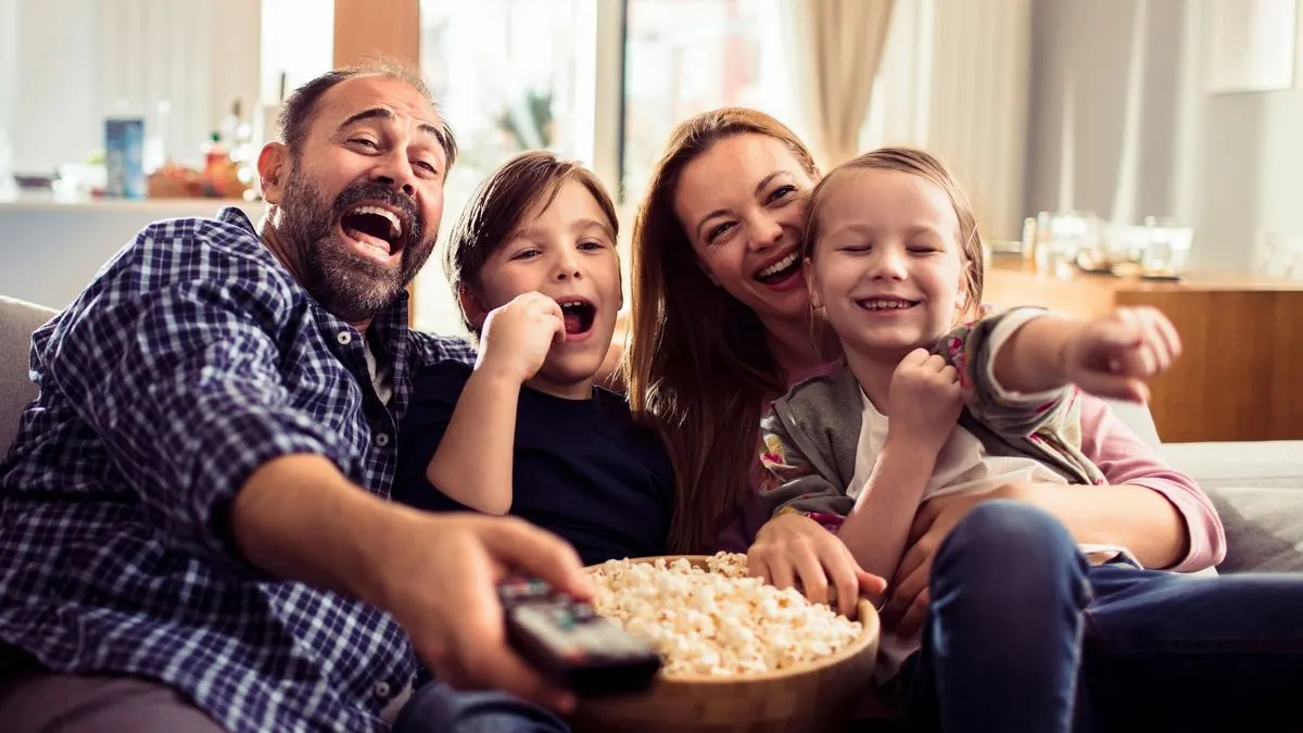 Familie sitzt gemeinsam auf dem Sofa, hält eine Schüssel Popcorn und schaut Fernsehen.