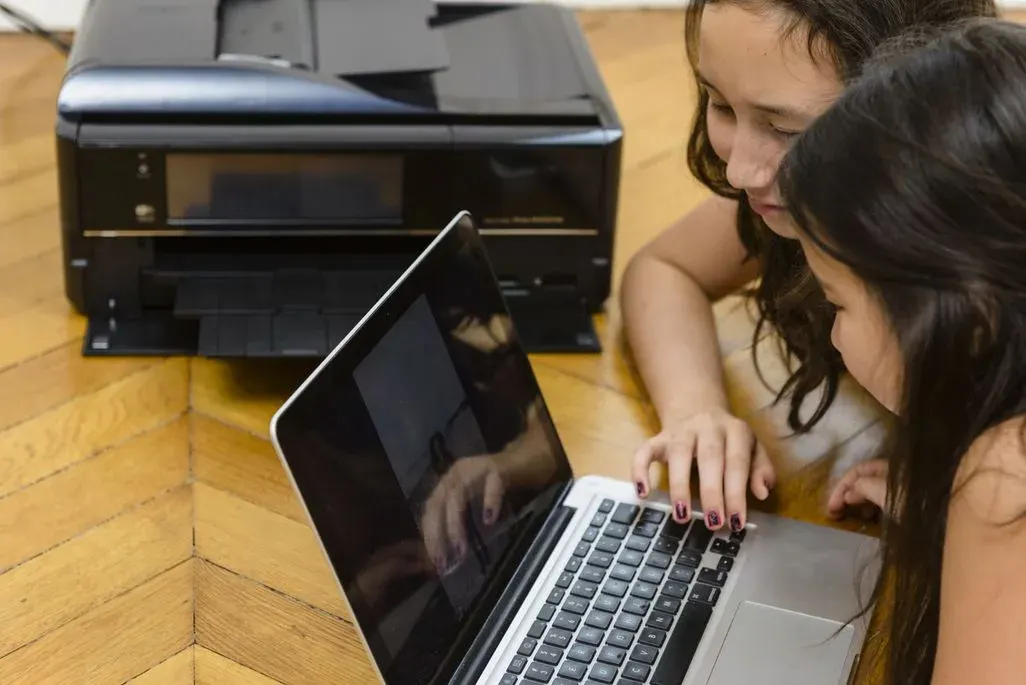 Zwei Frauen bedienen gemeinsam Laptop vor schwarzem Multifunktionsdrucker auf Holzboden.