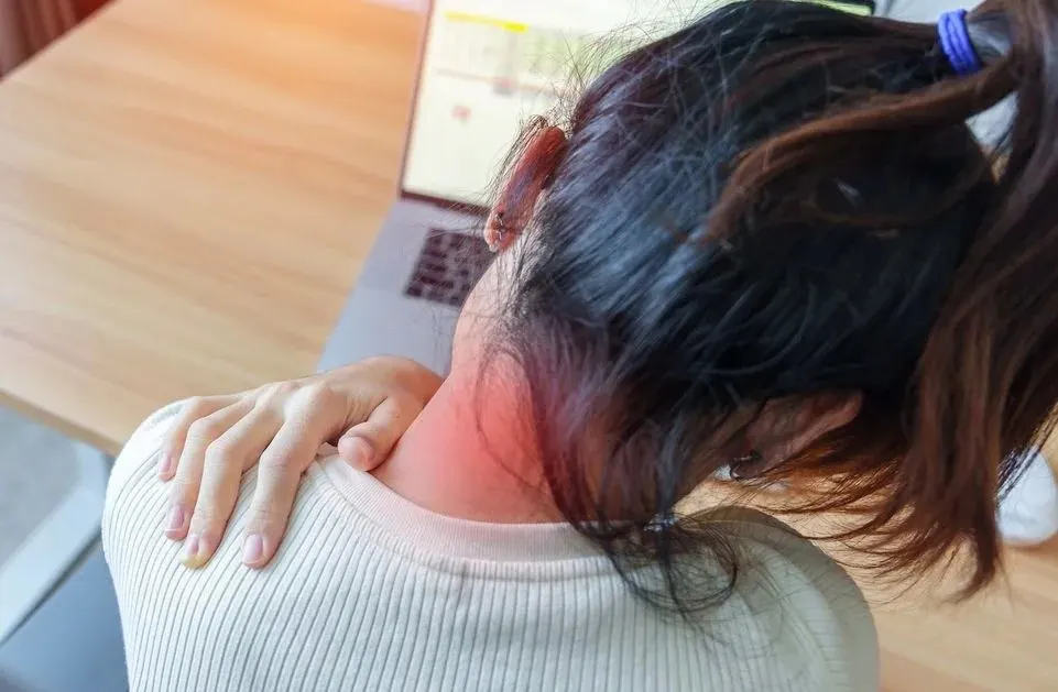 Eine Frau sitzt vor einem Laptop und hält sich die Schulter - Schmerzen werden durch eine rote Stelle am Nacken versinnbildlicht.