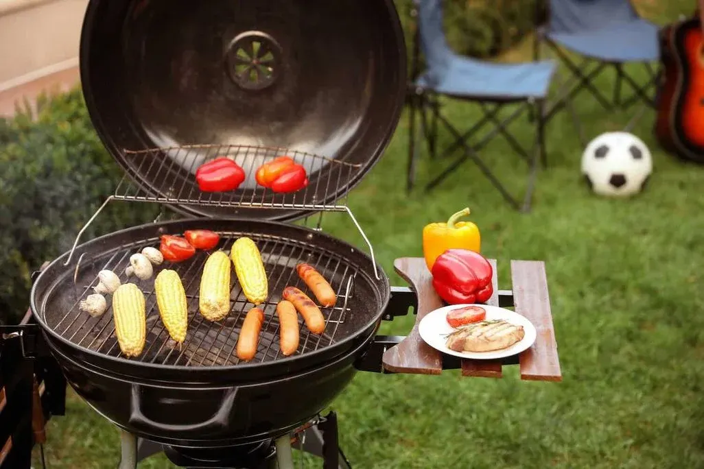 Ein schwarzer Kugelgrill im Freien mit aufgeklapptem Deckel. Auf dem unteren Rost grillen Maiskolben, Würstchen, Champignons und Tomaten, auf dem oberen Warmhalterost liegen rote und orange Paprika. Auf der seitlichen Holzablage stehen eine gelbe und eine rote Paprika sowie ein weißer Teller mit einem gegrillten Fleischstück und einer Tomate. Im grünen Hintergrund sind ein Fußball, ein Campingstuhl und eine Gitarre zu erkennen – typische Atmosphäre eines Gartengrillabends.
