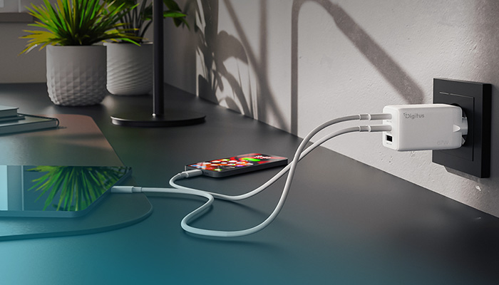 Ein weißes Digitus Multi-Port-Ladegerät steckt in einer schwarzen Wandsteckdose. Über zwei angeschlossene, weiße Kabel werden gleichzeitig ein Smartphone und ein Tablet aufgeladen, die auf einem Schreibtisch liegen.