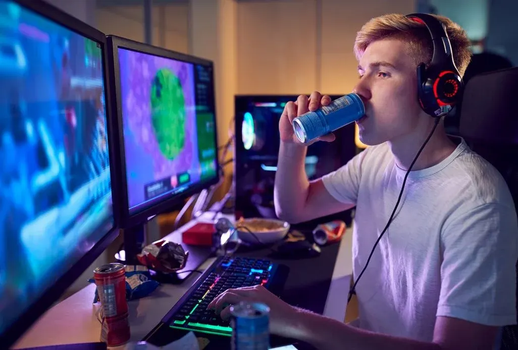Junger Mann mit roten Kopfhörern trinkt aus blauer Dose, spielt an beleuchteter Tastatur. Zwei Monitore zeigen Spiele, der Schreibtisch ist unordentlich.