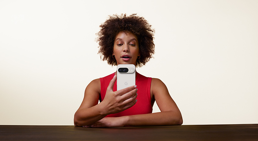 Eine Person hält ein weißes Smartphone mit einem Kameramodul vor einem hellen Hintergrund an einem dunklen Tisch.