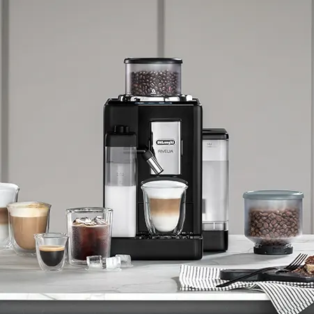 Schwarze Kaffeemaschine mit Kaffee, Milch und Eiswürfeln auf einem Tisch vor einem grauen Hintergrund.