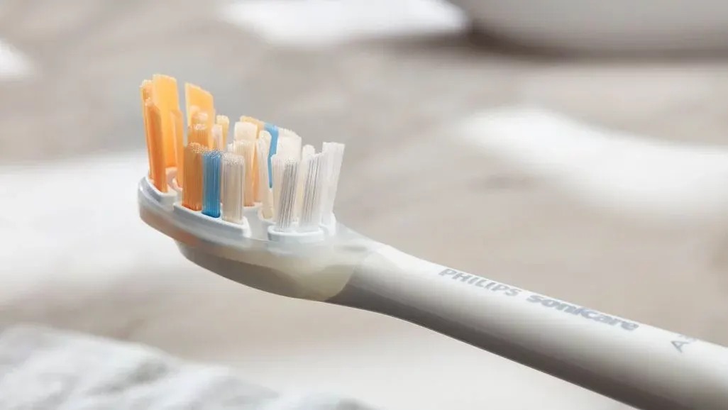 Eine Philips Sonicare elektrische Zahnbürste mit weißem Griff und Bürstenkopf mit mehrfarbigen Borsten (orange, blau und weiß), schräg auf unscharfem, hellem Hintergrund positioniert.