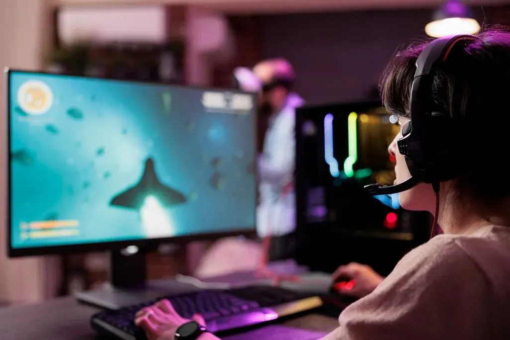 Frau mit Headset sitzt am RGB-beleuchteten Gaming-PC und steuert mit Tastatur und Maus ein Unterwasser‑Actionspiel, konzentriert vor großem Monitor.